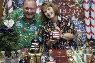 A smiling man and woman in Christmas shirts lean over a table of Gower Gin bottles in front of a North Pole backdrop, with a Small Drinks Producer of the Year award plaque