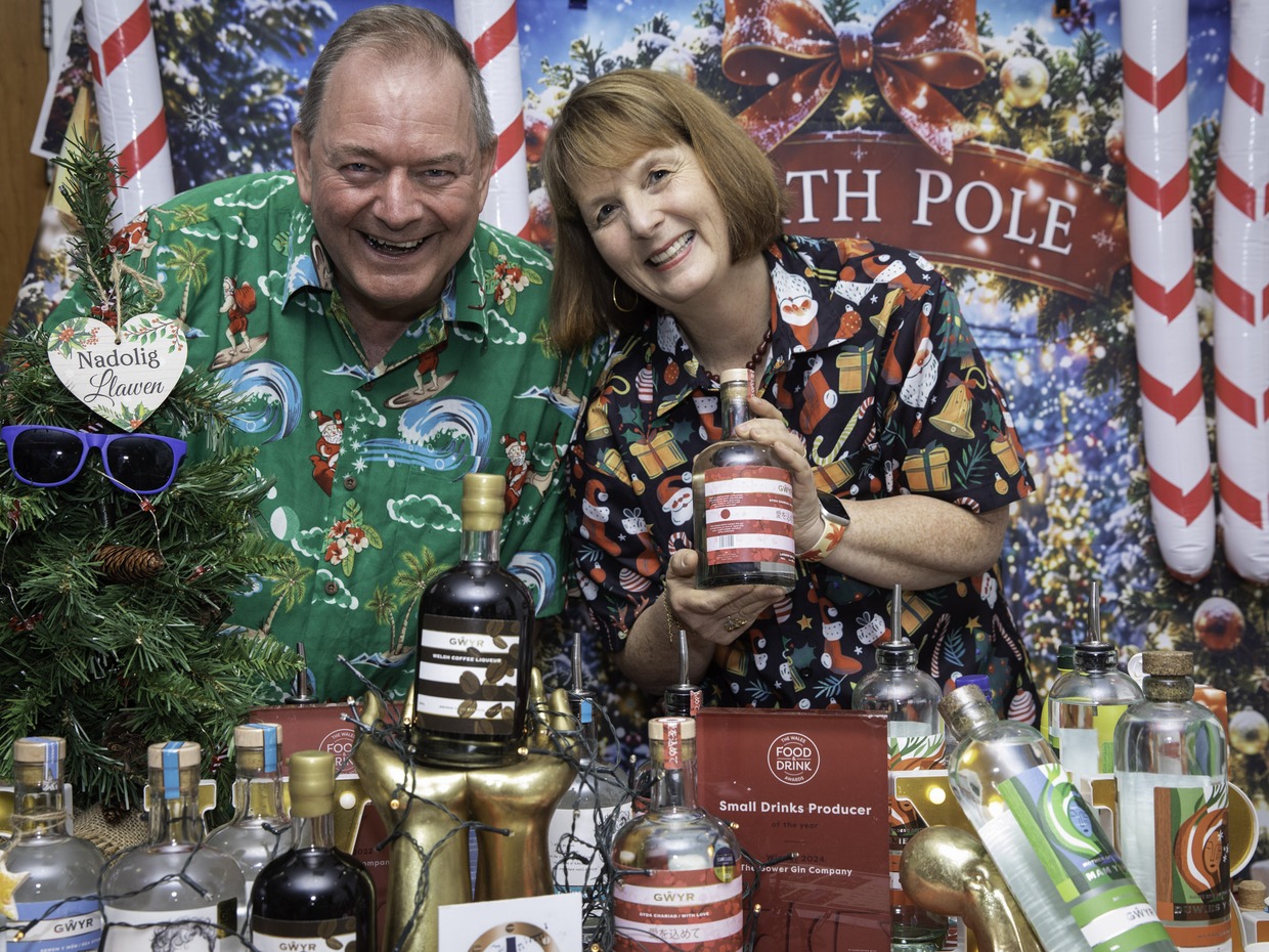 A smiling man and woman in Christmas shirts lean over a table of Gower Gin bottles in front of a North Pole backdrop, with a Small Drinks Producer of the Year award plaque
