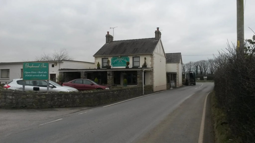 The Greyhound Inn while still open, a whitewashed pub with a green sign, stone porch and cars in the roadside car park