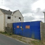 The former Greyhound Inn with its roof stripped to bare timbers, windows missing and a blue metal hoarding closing off the site beside a country road
