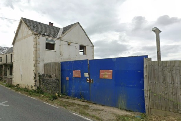The former Greyhound Inn with its roof stripped to bare timbers, windows missing and a blue metal hoarding closing off the site beside a country road