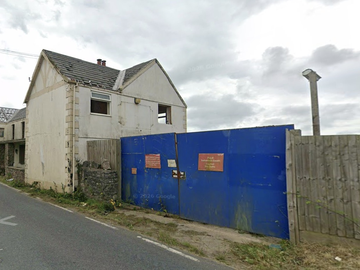 The former Greyhound Inn with its roof stripped to bare timbers, windows missing and a blue metal hoarding closing off the site beside a country road