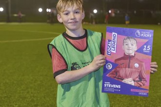 A young boy in a green bib on a floodlit football pitch holding up a Premier League Kicks Local Legend certificate with his own photo on it