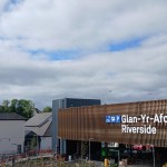 A multi-storey car park clad in bronze-coloured slats with a white illuminated sign reading Glan-Yr-Afon Riverside, with a ruined castle on the hill behind