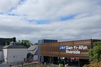 A multi-storey car park clad in bronze-coloured slats with a white illuminated sign reading Glan-Yr-Afon Riverside, with a ruined castle on the hill behind
