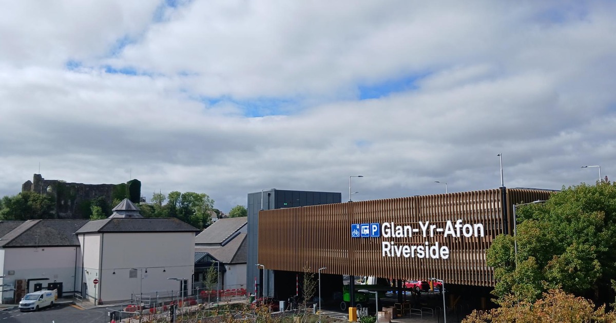 A multi-storey car park clad in bronze-coloured slats with a white illuminated sign reading Glan-Yr-Afon Riverside, with a ruined castle on the hill behind