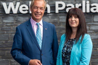 Nigel Farage in a blue suit and Helen Jenner in a turquoise jacket standing in front of the slate wall and Welsh Parliament sign at the Senedd