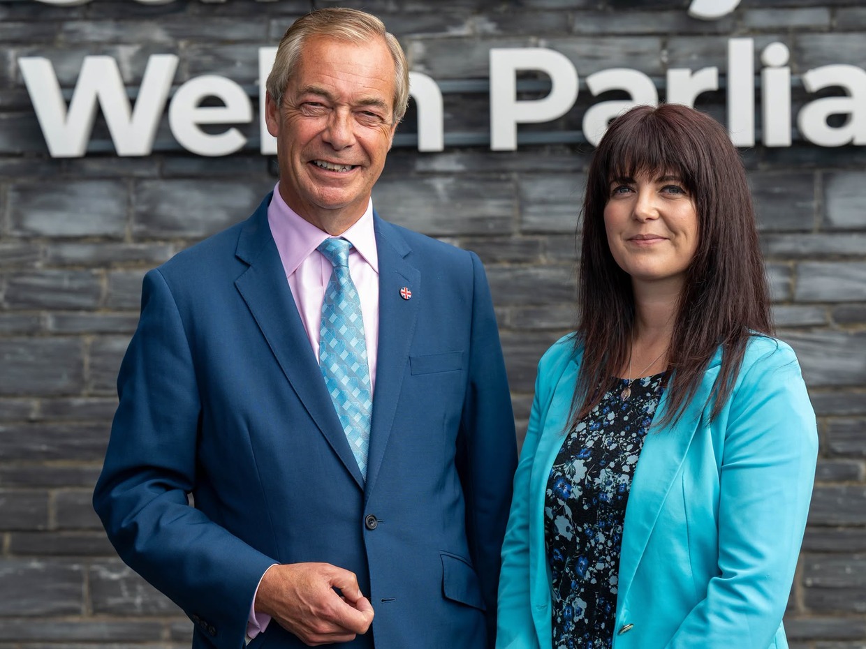 Nigel Farage in a blue suit and Helen Jenner in a turquoise jacket standing in front of the slate wall and Welsh Parliament sign at the Senedd