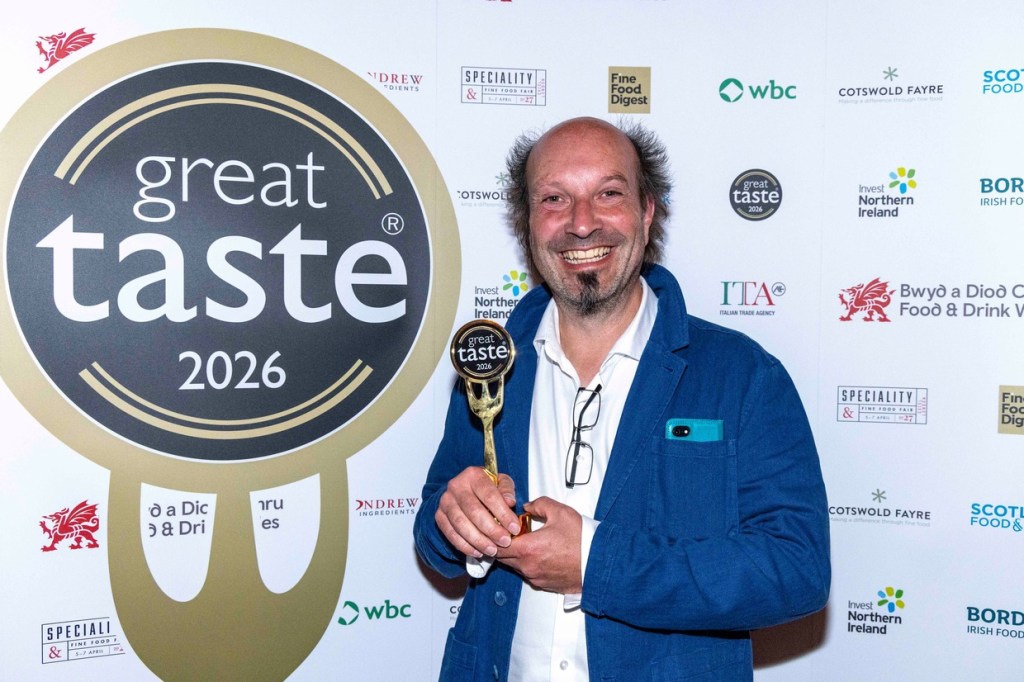 A smiling man in a blue jacket holding a gold fork-shaped Great Taste 2026 trophy in front of a sponsor board