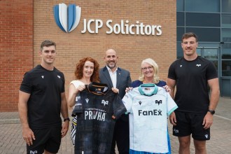 Two Ospreys players in black training kit flank three people holding up the club's black home and white away shirts outside a brick office with a JCP Solicitors sign