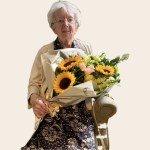 An elderly woman with white hair and glasses, in a patterned dress and cream cardigan, seated and holding a large bouquet of sunflowers and white flowers