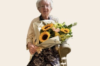 An elderly woman with white hair and glasses, in a patterned dress and cream cardigan, seated and holding a large bouquet of sunflowers and white flowers