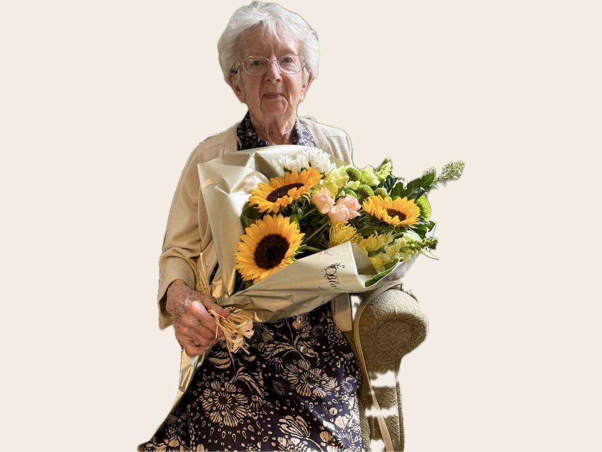 An elderly woman with white hair and glasses, in a patterned dress and cream cardigan, seated and holding a large bouquet of sunflowers and white flowers
