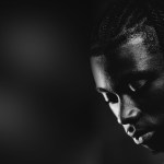 Black and white close-up portrait of Jeremy Monga looking down, his face half lit against a dark background
