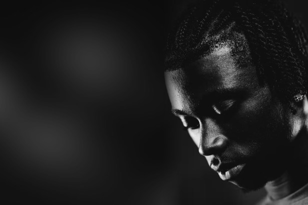 Black and white close-up portrait of Jeremy Monga looking down, his face half lit against a dark background