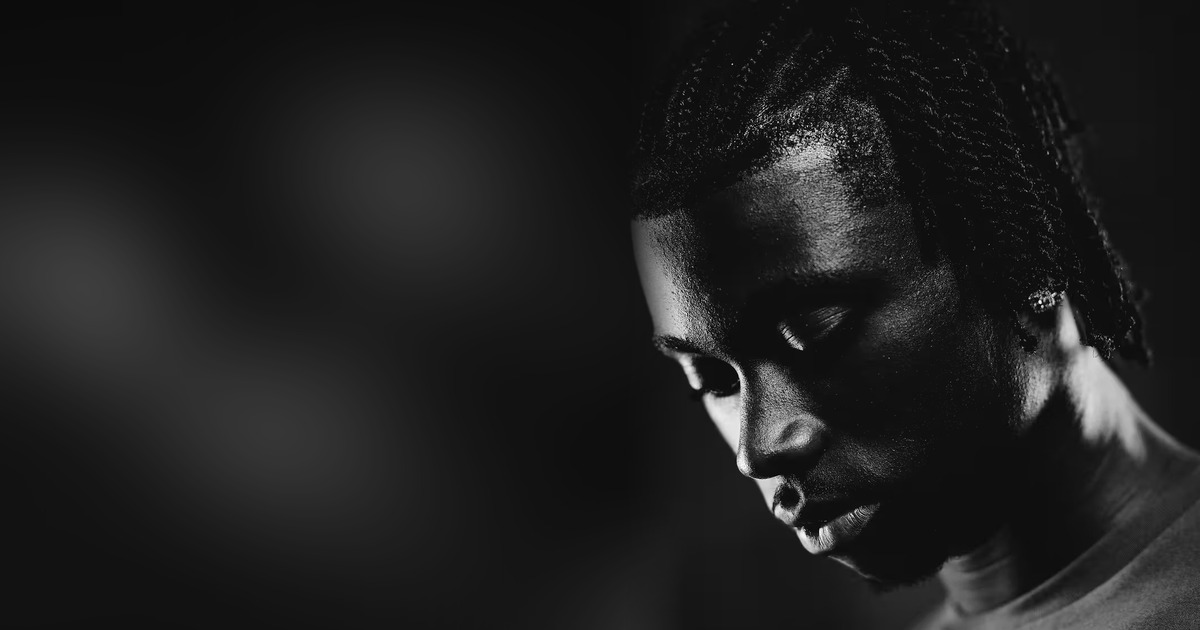 Black and white close-up portrait of Jeremy Monga looking down, his face half lit against a dark background