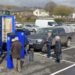A man paying at a blue pay-and-display machine in John Street car park, Carmarthen, with parked cars and houses on the hillside behind