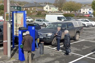 A man paying at a blue pay-and-display machine in John Street car park, Carmarthen, with parked cars and houses on the hillside behind