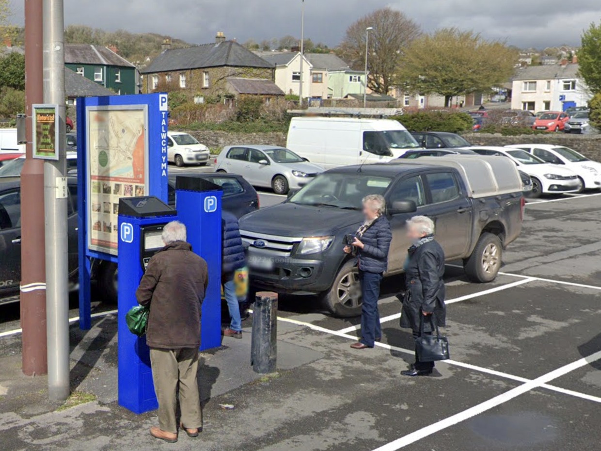 A man paying at a blue pay-and-display machine in John Street car park, Carmarthen, with parked cars and houses on the hillside behind