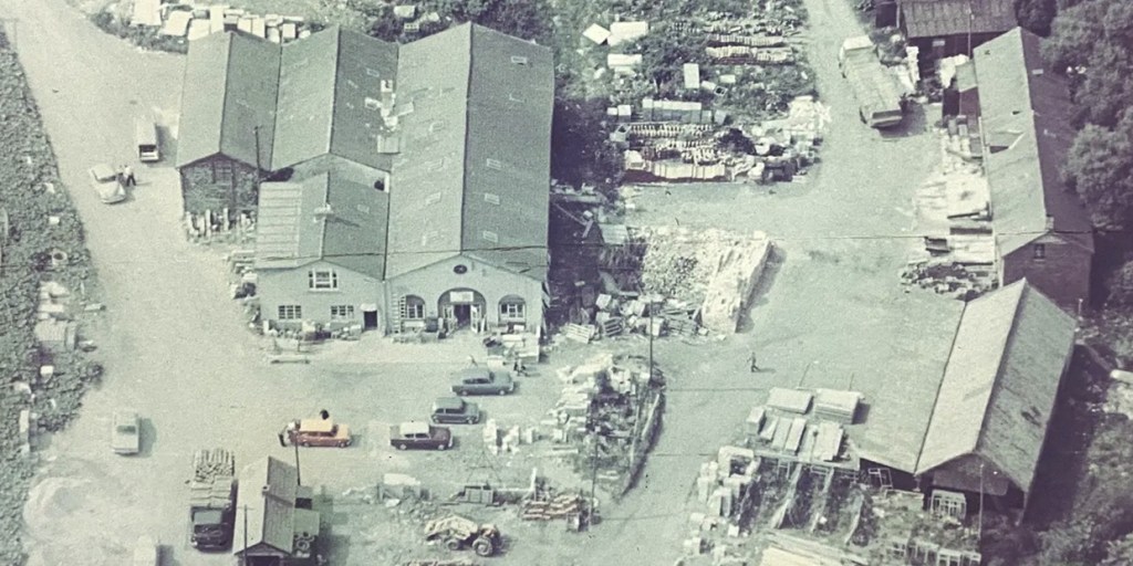 Faded archive aerial photograph of a timber and builders' yard with pitched-roof sheds, stacked materials and parked cars from the 1960s or 1970s