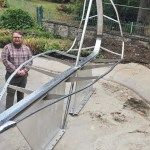 A man in a checked shirt stands beside the steel and glass Leaf Boat sculpture, mounted on plinths in a drained pond at Cwmdonkin Park, Swansea