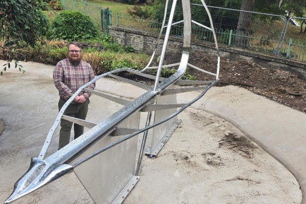 A man in a checked shirt stands beside the steel and glass Leaf Boat sculpture, mounted on plinths in a drained pond at Cwmdonkin Park, Swansea