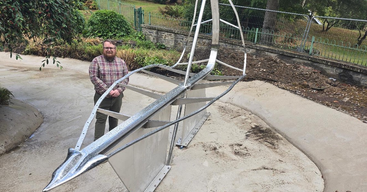 A man in a checked shirt stands beside the steel and glass Leaf Boat sculpture, mounted on plinths in a drained pond at Cwmdonkin Park, Swansea