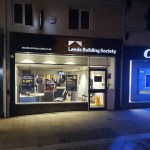 The Leeds Building Society branch at night, its lit window showing savings posters, between a Coral betting shop and another shopfront