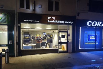 The Leeds Building Society branch at night, its lit window showing savings posters, between a Coral betting shop and another shopfront