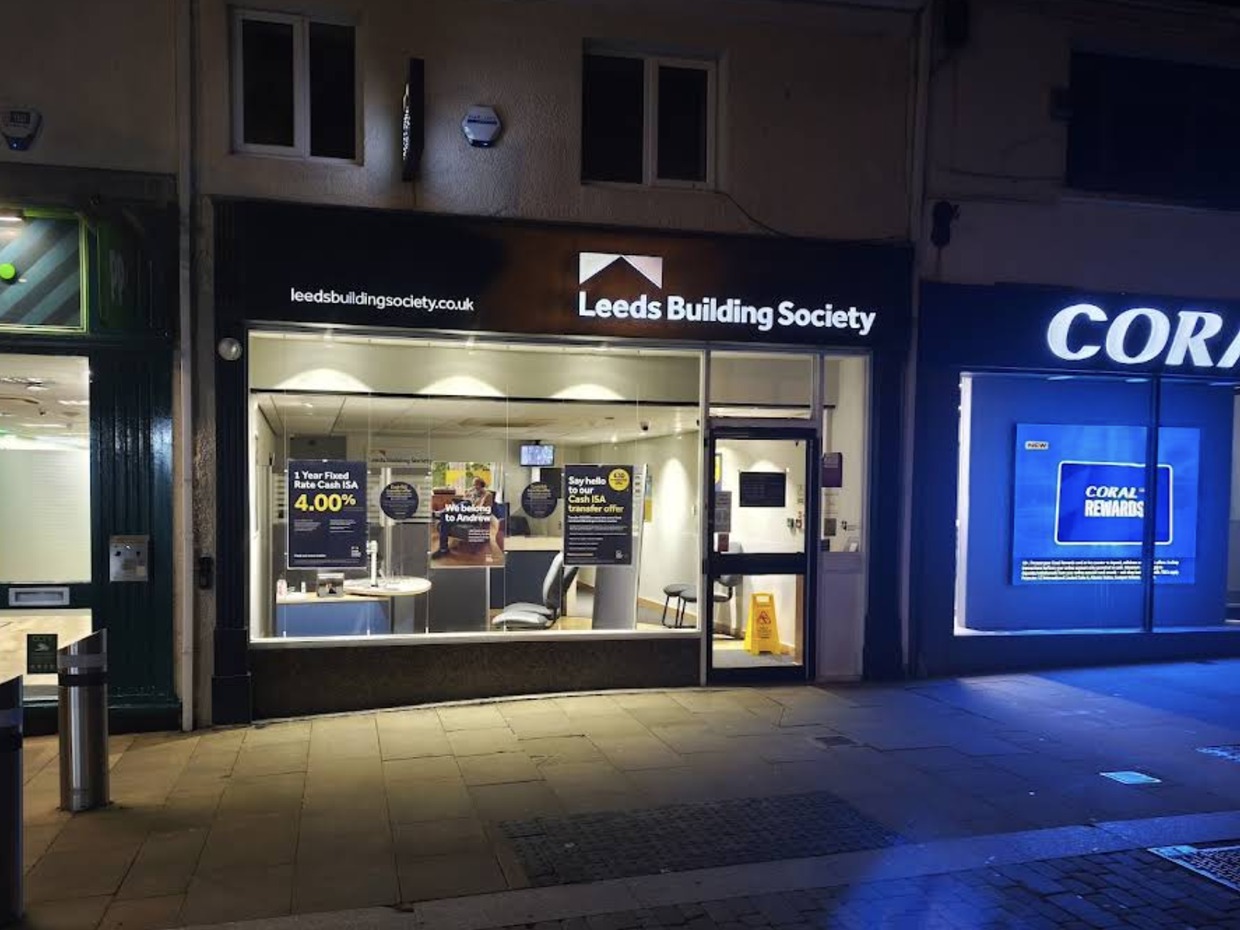 The Leeds Building Society branch at night, its lit window showing savings posters, between a Coral betting shop and another shopfront