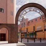 Architect's render of a tall rust-coloured steel arch reading Little Wind Street across the entrance to a brick-lined lane, with a smaller matching arch beside it and planters and cafe tables beyond
