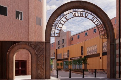 Architect's render of a tall rust-coloured steel arch reading Little Wind Street across the entrance to a brick-lined lane, with a smaller matching arch beside it and planters and cafe tables beyond