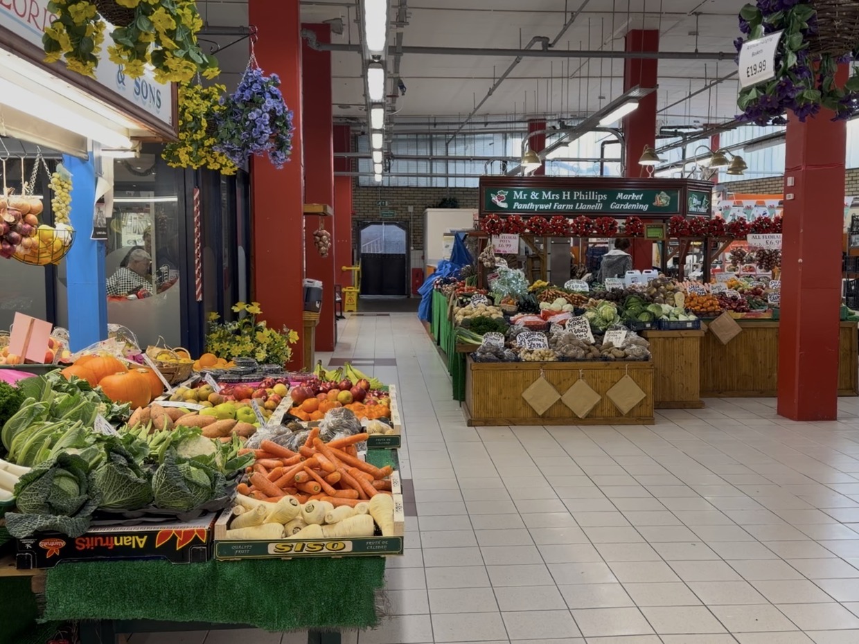 Fruit and vegetables displayed on the WD Seward and Sons stall inside Llanelli's indoor market, with produce stalls and a shopper along the aisle