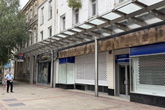 Shuttered shop units under a glass canopy on Stepney Street, Llanelli, with a To Let sign on the former bank beyond