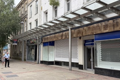 Shuttered shop units under a glass canopy on Stepney Street, Llanelli, with a To Let sign on the former bank beyond