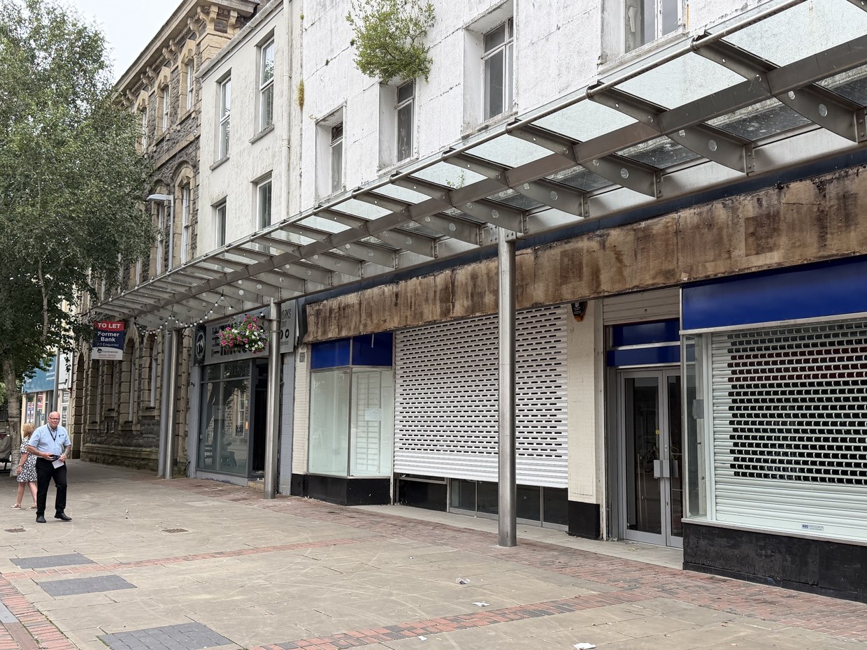 Shuttered shop units under a glass canopy on Stepney Street, Llanelli, with a To Let sign on the former bank beyond