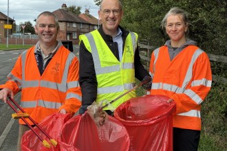Three people in hi-vis vests holding litter-pickers and red rubbish bags on a roadside verge, with houses behind