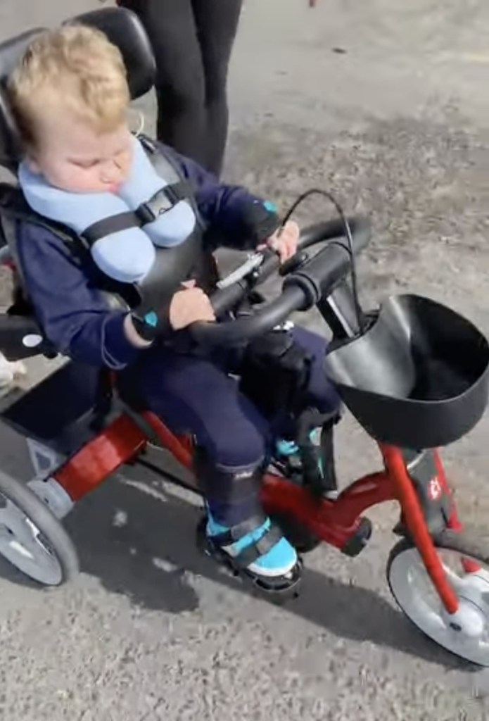 A three-year-old boy strapped into a red adapted tricycle with a padded head and neck support, on a gravel path
