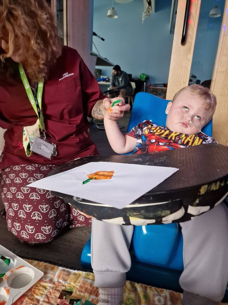 A three-year-old boy in a Spider-Man T-shirt sits in a blue chair with a tray, holding a paintbrush with help from a play practitioner