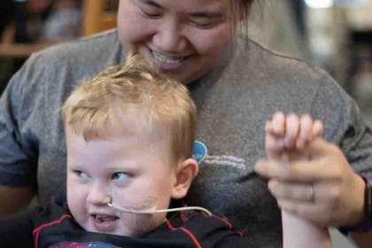 A smiling three-year-old boy with a feeding tube holds his hand up high, supported by a laughing physiotherapist behind him