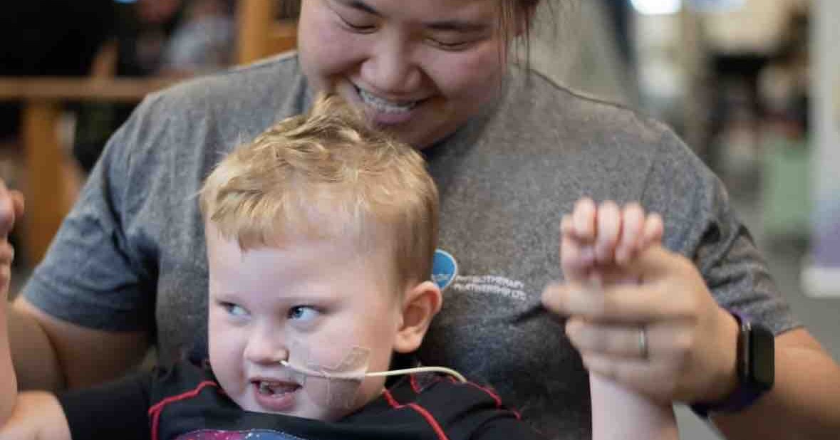 A smiling three-year-old boy with a feeding tube holds his hand up high, supported by a laughing physiotherapist behind him