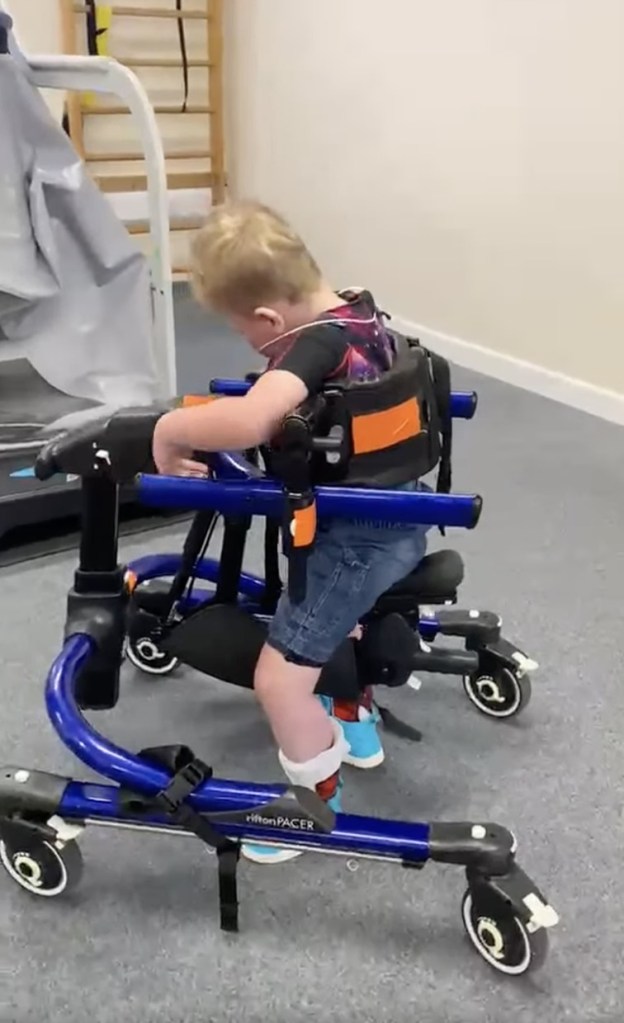 A three-year-old boy takes a step in a blue Rifton Pacer walking frame in a physiotherapy gym
