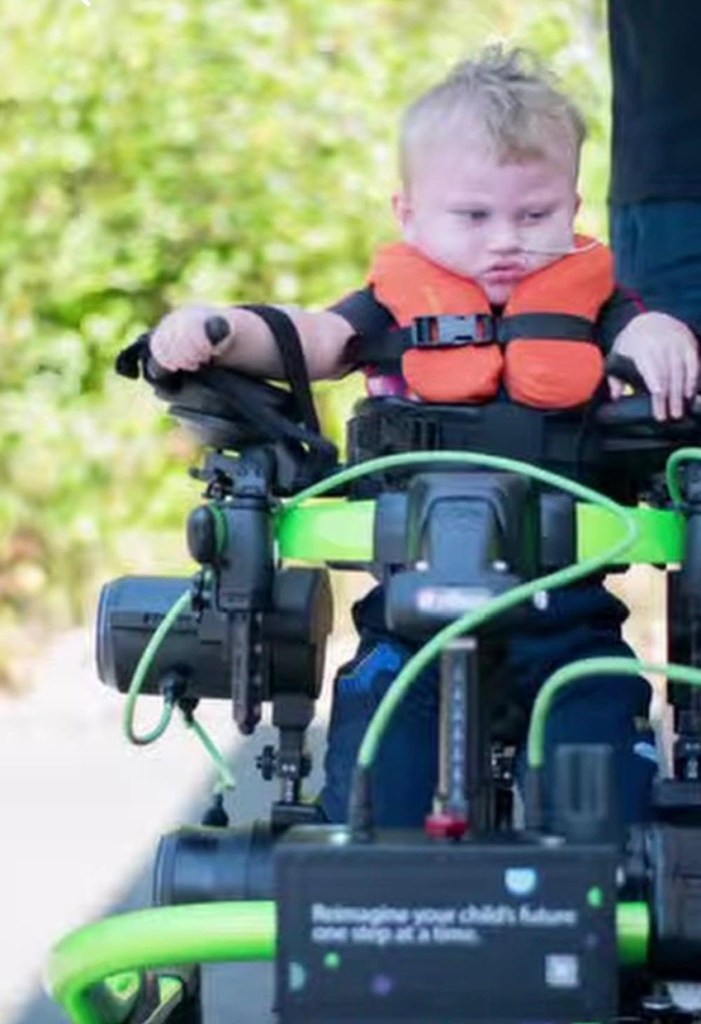 A three-year-old boy in an orange support vest stands in a green Trexo robotic walking frame outdoors