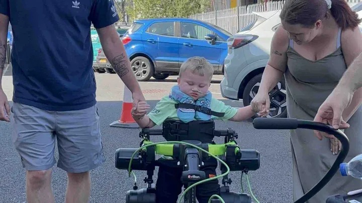 A three-year-old boy stands in a green Trexo robotic walking frame in a car park, holding the hands of two adults either side of him