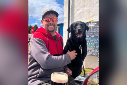 Luke King smiling in sunglasses and a cap with his black Labrador Digger
