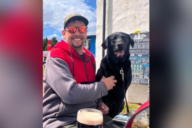 Luke King smiling in sunglasses and a cap with his black Labrador Digger