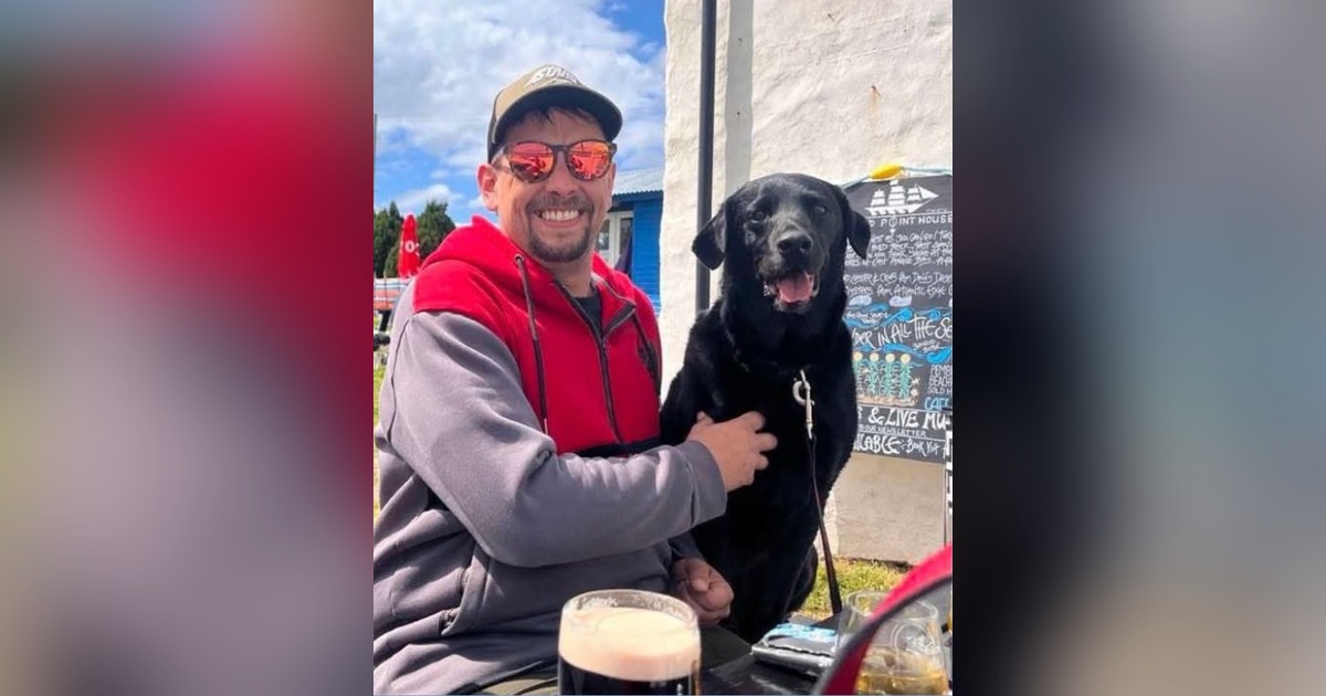 Luke King smiling in sunglasses and a cap with his black Labrador Digger