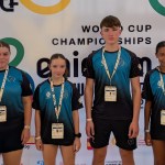 Four young taekwon-do athletes in matching blue and black club shirts with accreditation lanyards, standing in front of the World Cup Championships banner in Benidorm