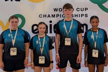 Four young taekwon-do athletes in matching blue and black club shirts with accreditation lanyards, standing in front of the World Cup Championships banner in Benidorm