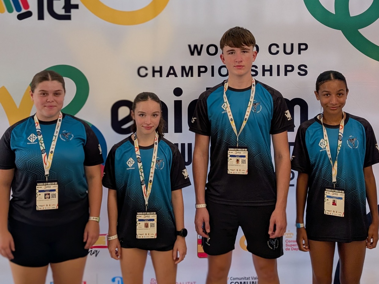 Four young taekwon-do athletes in matching blue and black club shirts with accreditation lanyards, standing in front of the World Cup Championships banner in Benidorm